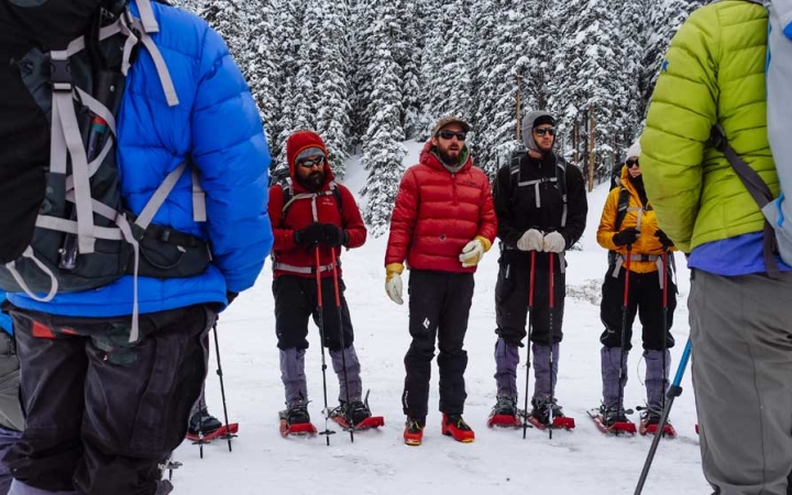 a group of veterans wearing snow gear and snowshoes stand in a circle while an instructor speaks